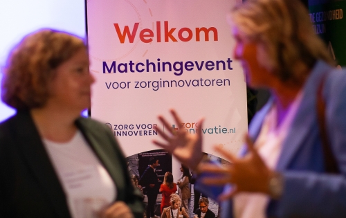 De roll-up banner van het Matchingevent, waarop staat 'Welkom - Matchingevent voor zorginnovatoren'. Daarvoor staan twee vrouwen die in gesprek zijn. Focus van de foto ligt op de banner. 