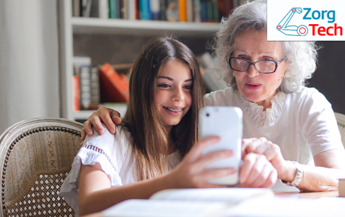 Oudere vrouw en jong meisje zitten naast elkaar aan tafel en kijken samen naar een iPhone scherm. 